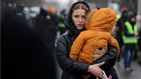 A woman (in black coat with hood) carries a child (wearing orange coat)