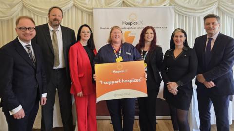 Four women and three men in suits are standing around a woman with a orange sign which reads "I support hospices".