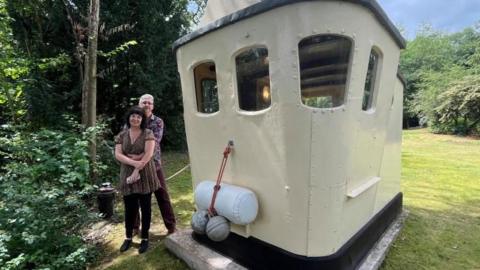 Wide-angle lens photograph of Richard and Abi stood next to their cabin - a wooden and steel structure painted cream with two small windows on each side. On the front of the cabin are buoys and fenders, and it's 'tied up' to a black mooring post. It's in a garden, with green trees and bushes along the edges of green grass. Richard and Abi are stood with their arms around each other. 