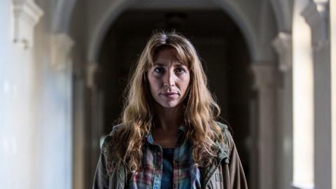 Daisy Haggard as Mira Matteson. She is weating blue check shirt with a green coat
