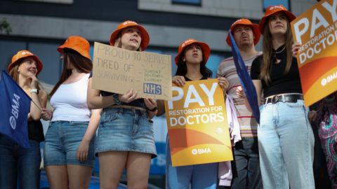 Six people taking part in strike action. They are carrying placards and wearing orange BMA hats. 