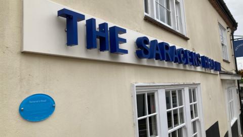 Cream-coloured wall of pub with "The Saracens Head" in large blue letters above the windows. A blue plaque is attached to the wall