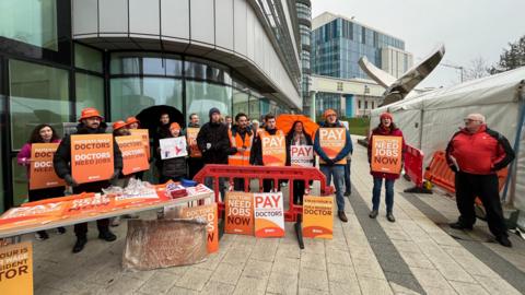 A group of people standing outside a glass building holding orange cards. They say things like "patients need doctors, doctors need jobs" and "doctors need jobs now". They are wearing orange bucket hats and beanies, and have an orange table with more signs leaning against it.