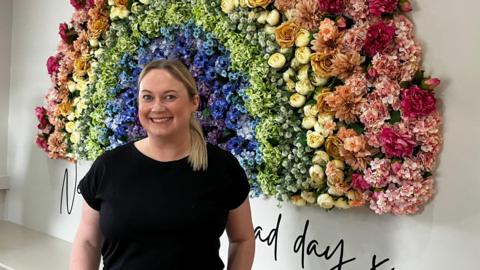 A woman smiles at the camera. She is wearing black. She is standing in front of a rainbow made of flowers on a white wall.