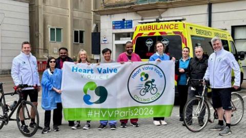 Two cyclists standing for a photo outside a building with people cheering them on stood between them. An ambulance is behind them.