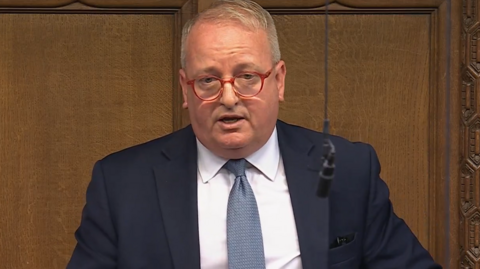 A man with short fair hair and red-framed glasses stands in the House of Commons in a navy blue suit, white shirt and blue tie. The wood panelling of the chamber is behind him, and a microphone hangs on a wire in front of him.