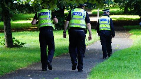 Three uniformed police officers wearing high-viz vests are walking down a path lined with trees and grass.