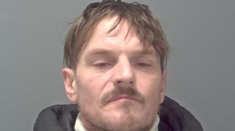 A mugshot of a man wearing a hooded jacket over a white rollneck jumper. He is standing in front of a grey background and looking into the camera.