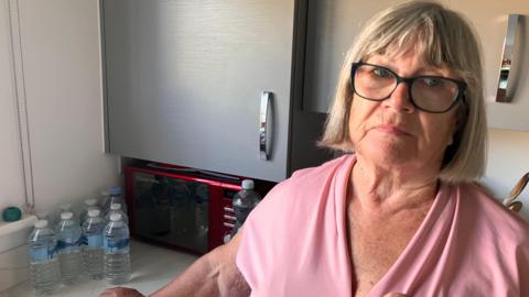 A woman with glasses and a pink top in her kitchen with a cluster of 10 plastic bottles of water on her work top, in front of a microwave.