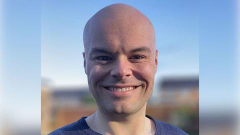 Ben smiles at the camera in a close-up picture against a blue background. 
