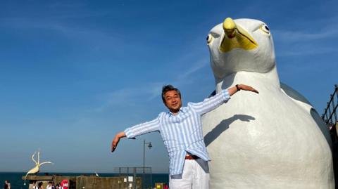 Hiroshi Suzuki is wearing a stripy blue shirt, and has his arms outstretched as if mimicking a bird. He is smiling and there is massive sculpture of a white seagull with a yellow beak directly behind him.