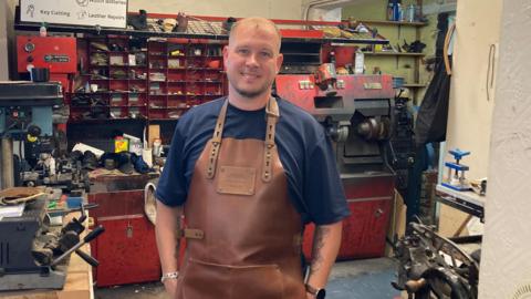 A photo showing a man with short hair standing in a workshop. He is wearing a blue T-shirt and a brown, leather apron. In the background are red cubby holes, a large shoe repairing machine and various cobbler's tools.