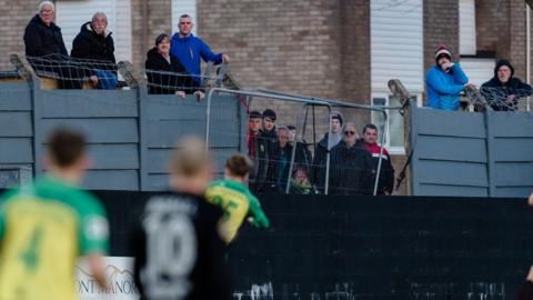 Caernarfon fans watching their team face Connah's Quay from behind the wall at the club's Oval ground