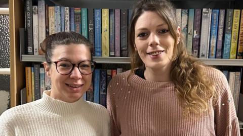 Two women, one with hair tied back and wearing glasses, the other with long brown hair, stand in front of book shelves