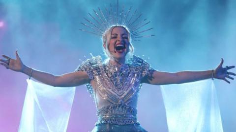 A singer with an elaborate wired headpiece and floaty material coming from her arms sings out with her arms outstretched