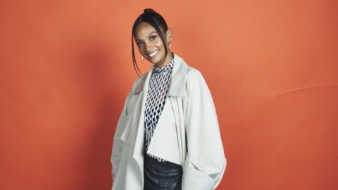 A woman smiling stood against a red background. Her hair is tied back only leaving to sections in the front which frames her face. She is wearing a white mesh shirt and leather bottoms with an oversized cream jacket on top.