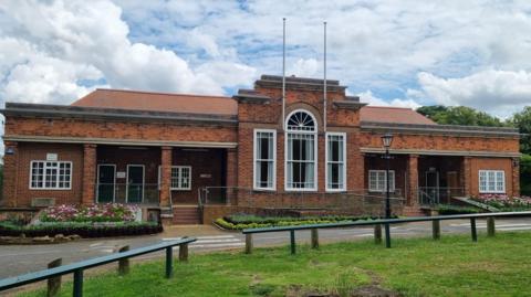 The outside of a red-brick pavilion, with two flag poles, several white windows and flower beds outside, with railings around the building. A grass area is in front of the building. 