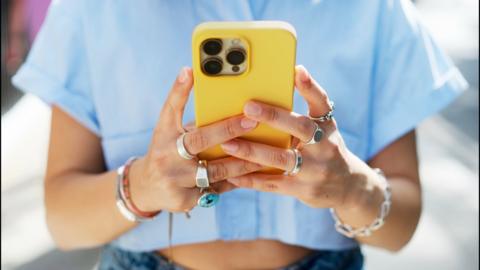 Mid view of someone with lots of rings holding a yellow phone in front of them.