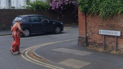 A woman dressed in a long red dress is crossing Frederick Road at its junction with St James Road, on which a blue car is being driven. The sign for Frederick Road is showing on the right of the shot. 