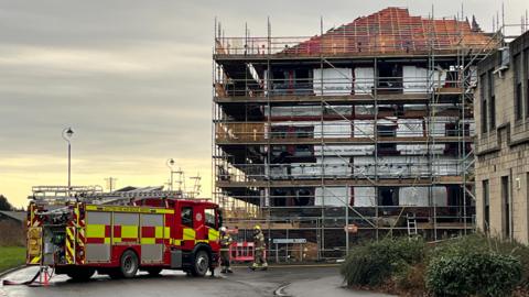 Daylight image of a three story block of flats - looking to be still under construction - with scaffolding surrounding it and a fire appliance and firefighters standing outside.