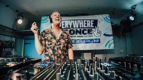 DJ Fatboy Slim stands behind a mixing console inside a low-ceiling venue, wearing headphones and a patterned shirt, with turntables, sliders and dials in the foreground and a large Everywhere At Once banner on the back wall under stage lighting.