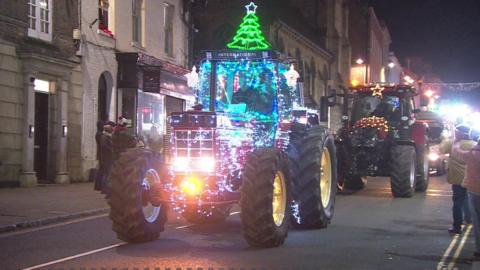 A row of festive tractors driving down a street at night. They are covered with brightly coloured Christmas lights and people are watching from the pavement wearing hats and coats.