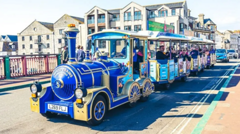 An image of a colourful land train with visitors on board going across a bridge in Weymouth