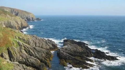 A coastline of rocky cliffs with green grass atop them, and blue sea stretching off into horizon on the right side of the image. 
There is a large rocky outcrop at the bottom of the cliffs. 