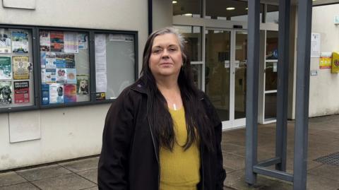 Woman with yellow jumper and black jacket stood in front of a building and looking at a camera. Behind her, on the wall of the building, are three noticeboards with several posters on them