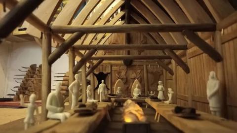 A model of the interior of a viking longhouse with model figurines sitting and standing either side of a long dining table.