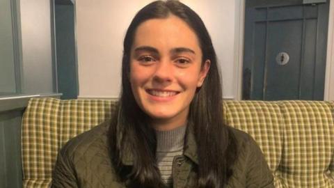 Jenny Hall. She has long brown hair and is smiling at the camera. She is wearing a green coat and sitting on a sofa.