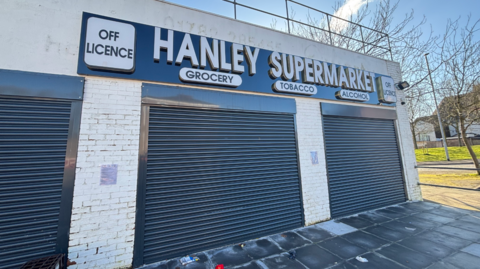 A low-roofed white brick building with shutters down. A sign on the front reads: "Hanley Supermarket - off-licence - grocery - tobacco - alcohol".