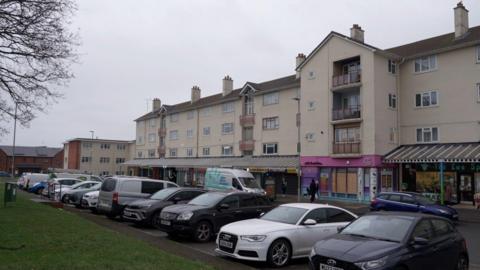 A row of businesses with three storey flats above. There is a pavement and a road outside the premises. There is a row of cars parked on the opposite side of the road in parking bays. The sky is grey and overcast.