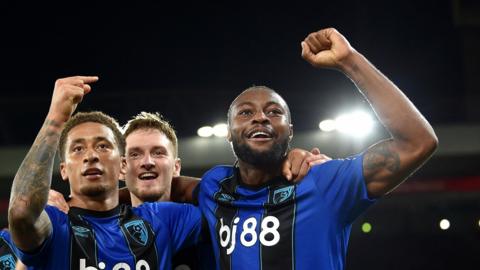 Antoine Semenyo celebrates a Bournemouth goal by raising his left arm up, bent at the elbow, above his head, while two team-mates join him, with one pointing at Semenyo. All are wearing blue and black vertical striped kit