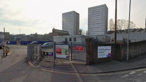 A Google StreetView image showing the entrance to the Hollingdean Depot which has large metal gates and fences and a council sign on the front with the name of the depot on it. In the background are two tower blocks.