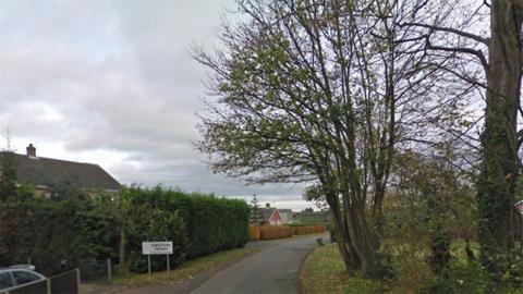 East Lound Road as it enters Owston Ferry with a sign for the town and hedges and trees on either side