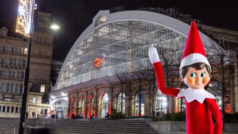 Liverpool Lime Street train station entrance sits on top of steps, its lit up as it is night time and there's a black lamp post to the left of the picture which has a lighted Christmas decoration. To the right of the picture is a super imposed toy elf waving