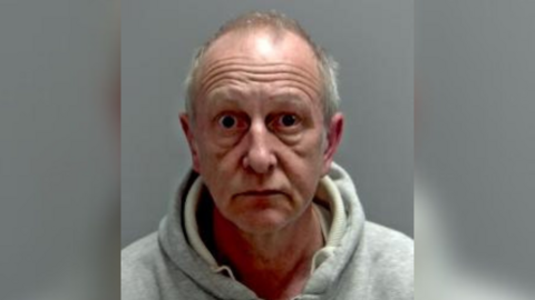 A custody mugshot taken of Martin Pearson, who is wearing a grey hoodie and has grey hair. He is wide-eyed.