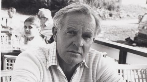 A black and white photo of a man wearing a long-sleeved pale top with the sleeves rolled up. He's sitting at a table and leaning forward on his elbows, and looking, unsmiling, at the camera. There's a wooden railing and some other people sitting at tables behind him.
