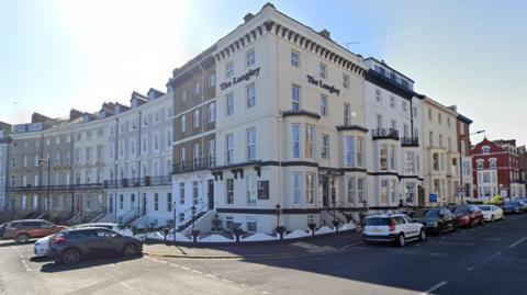 The Langley Hotel catches the eye on the corner of the street with its white walls, black decor, and its name in black on the front and side. There are cars parked outside and a blue sky above.