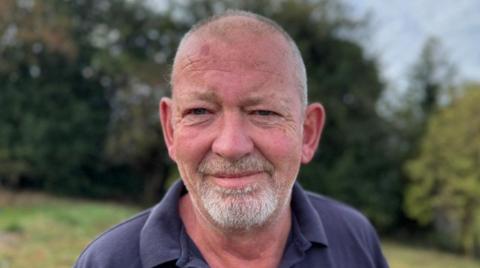A man with a white beard is wearing a blue top. Grass behind him is blurred.