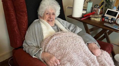 Margaret Linehan is seen with white, curly hair siting in a red armchair, smiling gently. She is wrapped in a blanket and wears a light grey cardigan over a darker top. 