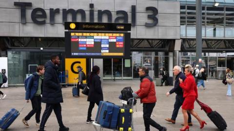 People lug suitcases in the plaza outside Terminal 3 at Heathrow. An information board shows the various airlines and their corresponding check-in zones.