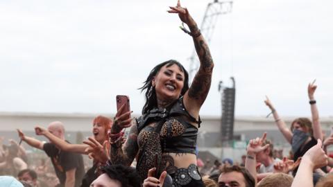 A woman stood on the shoulders of a man with her left hand in the air with other people behind her also standing on people's shoulders enjoying music at the Reading Festival 2025.
