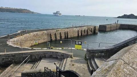 A wide shot of the West Hoe pier. It is fenced off. In the background is the Plymouth Sound and a cruise ship.