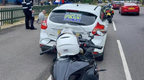 A sign reading "No Insurance, seized by police" at the back of a car. A police offer is seen nearby with a citizen who sits on the pavement. Other people are near them. Cars keep driving on the road.