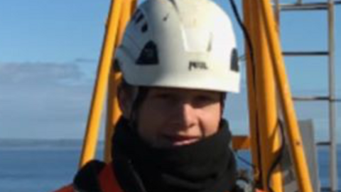 A man with a white helmet and black scarf or snood, stands and smiles while working at a construction site.