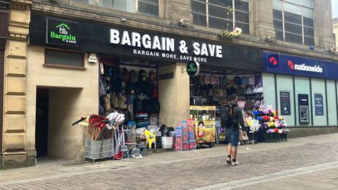 A shop called 'Bargain and Save' which is in an historic building but has lots of items outside like brooms and plastic boxes and flowers. There is a nationwide bank next door.