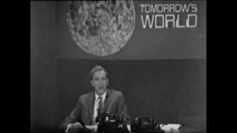 Tomorrow's World presenter sits at a desk with phones, below a sign with the Moon and 'Tomorrow's World' on it