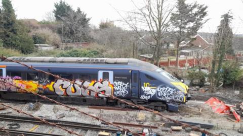 A blue Southeastern high speed train covered in multi-coloured graffiti being put back on the tracks. There are trees and a number of properties in the background. 